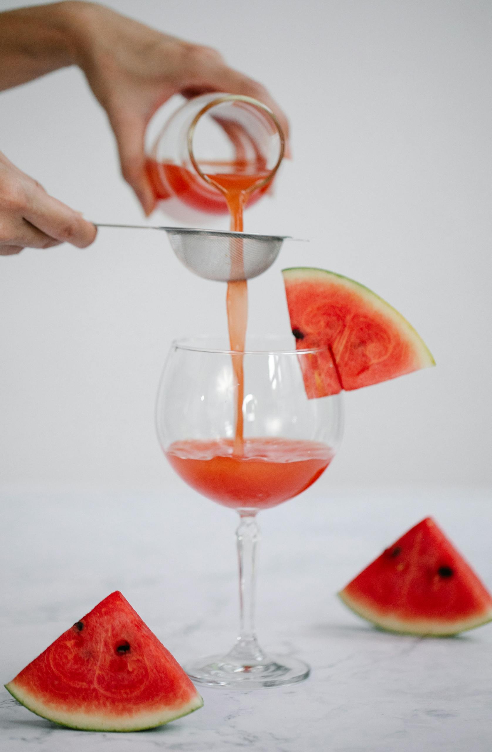 Refreshing watermelon juice being poured through a strainer into a wine glass with watermelon slices.