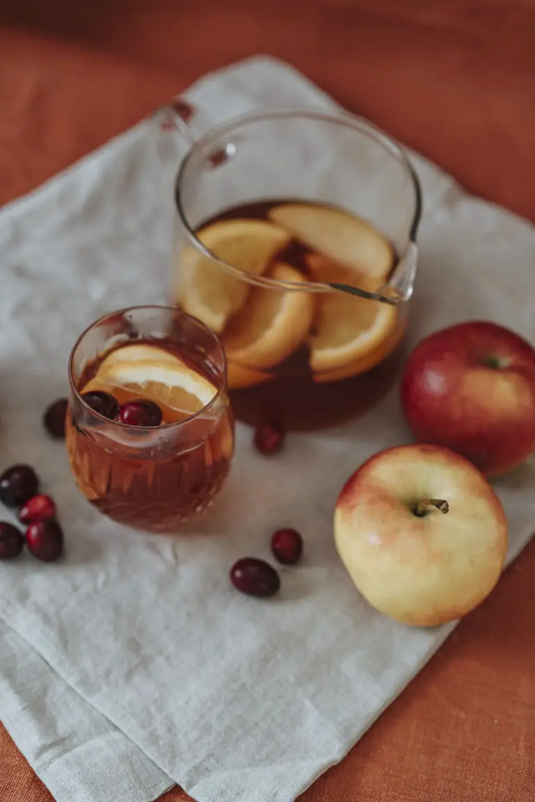 Warm autumn beverage with apples, cranberries, and orange slices in a glass pitcher and cup.